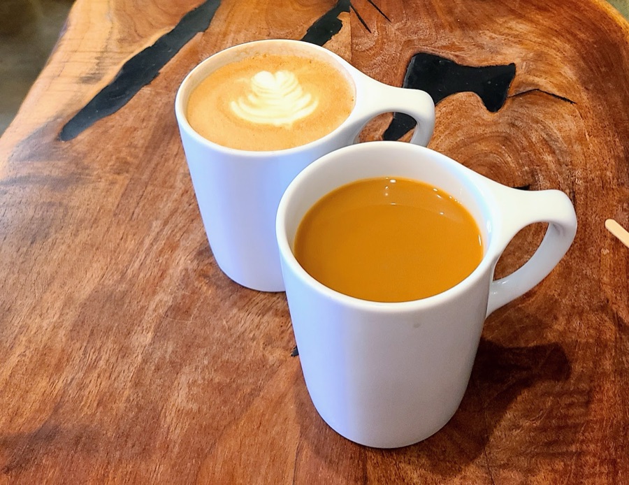 A latte with leaf art beside a cup of espresso on a live-edge wood table.